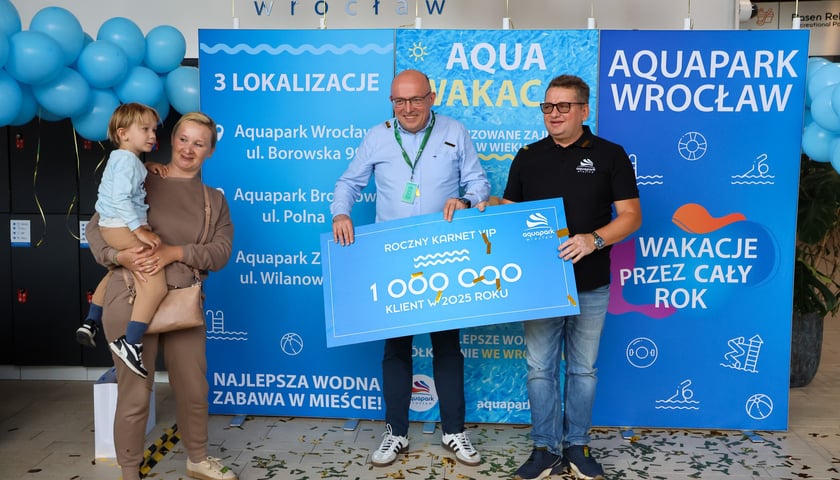 Ewelina Kowalenko z synem Julianem odebrali całoroczny karnet dla milionowego gościa Aquapark Wrocław w 2025 r. Na zdjęciu kobieta z dzieckiem na ręku, dwóch mężczyzn trzyma karton w formie karnetu