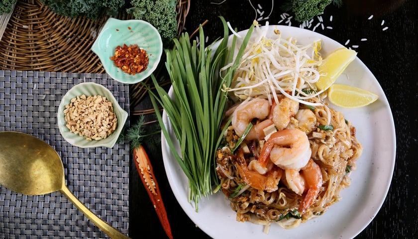 Po lewej stronie dwie miseczki: jedna z orzeszkami ziemnymi, druga z chilli, obok talerz z tajską potrawą z krewetkami. 