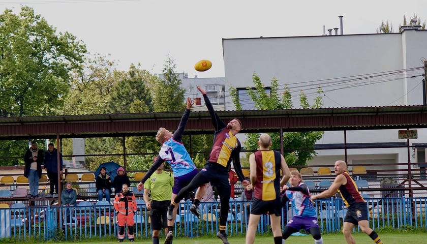 Zawodnicy w czasie meczu futbolu australijskiego w wyskoku walczą o lecącą w powietrzu jajowatą piłkę.