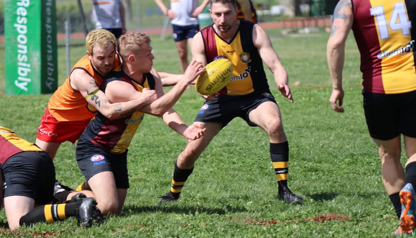 Ostre starcie kilku graczy na trawiastym boisku w czasie walki o jajowatą piłkę podczas meczu futbolu australijskiego.