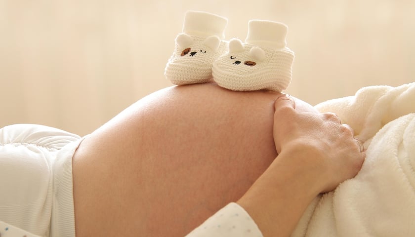 Ciężarna kobieta leżąca na plecach a na jej brzuchu leżą dziecięce buciki.