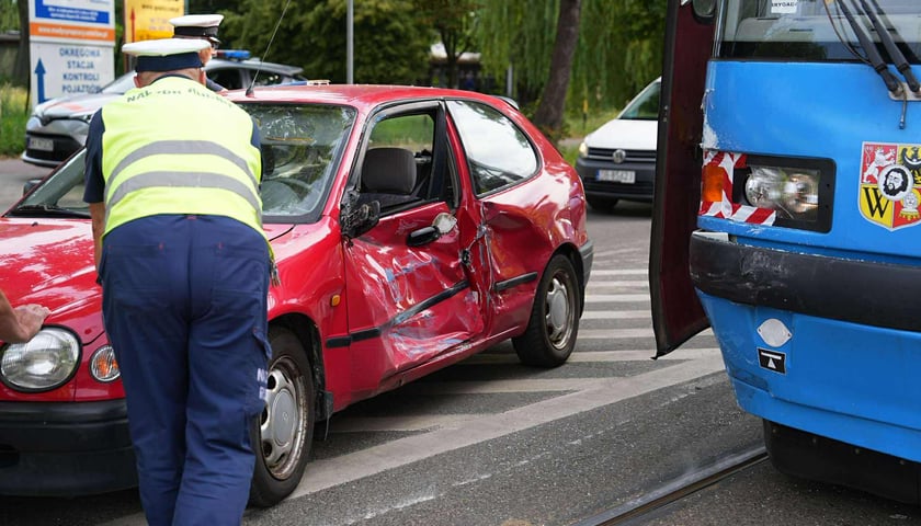 Czerwony samochód z pogniecionymi drzwiami po zderzeniu ze stojącym obok tramwajem, pracownicy nadzoru ruchu