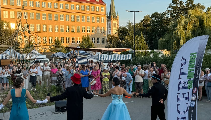 Czworo artystów Operetki Wrocławskiej: dwie kobiety w długich niebieskich sukniach i dwóch mężczyzn we frakach i cylindrach sfotografowani tyłem, bo stoją przodem do publiczności zgromadzonej na podwórku jednego z osiedli Wrocławia