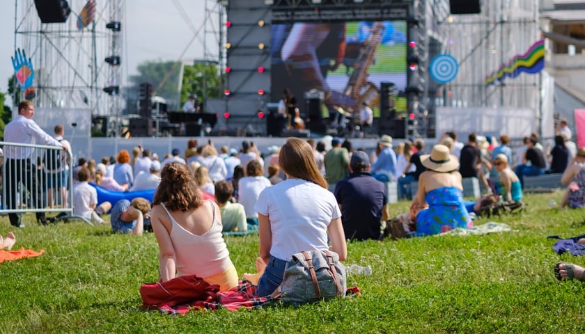 Koncert plenerowy. Zdjęcie ilustracyjne. Na zdjęciu widać ludzi, którzy siedzą na trawie przed duża sceną