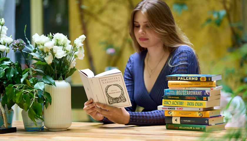 Młoda kobieta czyta książkę pt. „Robinson Cruzoe” wydaną w serii Biblioteka Narodowa, obok stos książek