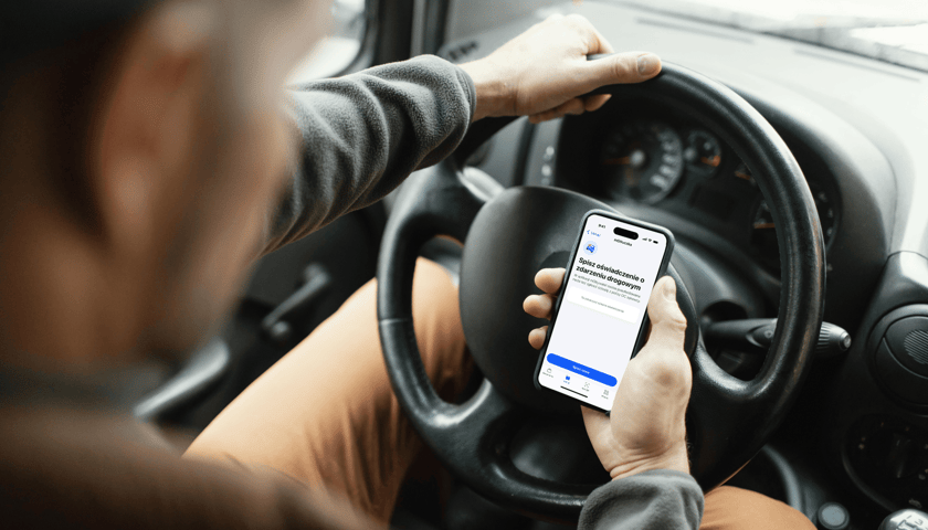 Mężczyzna trzyma w ręku kierownicę samochodu i telefon na którym widać aplikację mStłuczka