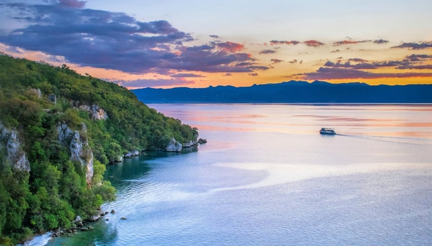 Skalisty brzeg jeziora porośnięty drzewami z widokiem na przepływający statek i odległe pasmo górskie. Jezioro Ochrydzkie w Macedonii Północnej