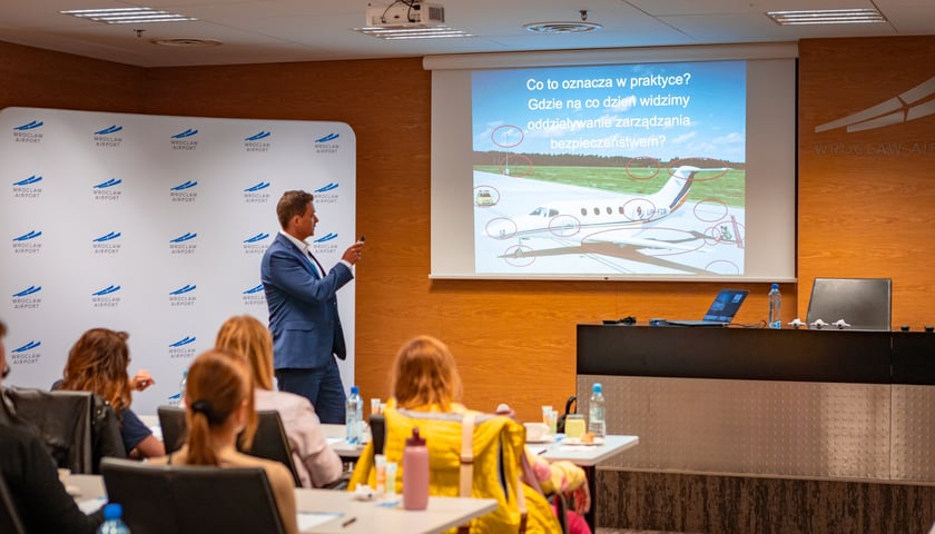 Mężczyzna w garniturze przy ekranie omawiający prezentację oraz osoby siedzące w ławkach i słuchające szkolenia. Warsztaty dla osób bojących się latać samolotem zorganizowane przez Port Lotniczy Wrocław.