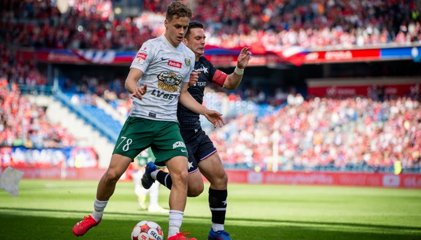 Dwóch piłkarzy walczy o piłkę na boisku na tle trybun pełnych kibiców. Tommaso Guercio ze Śląska Wrocław podczas mecz