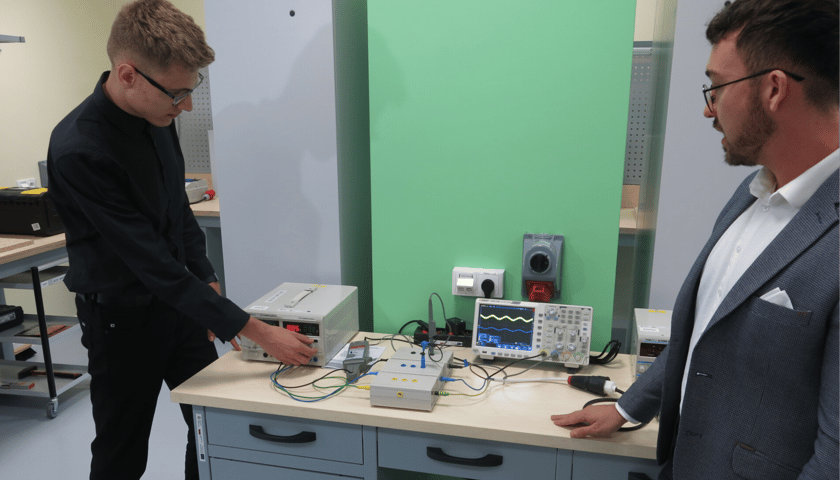 Uczeń i instruktor przy stole na którym znajdują się urządzenia elektryczne. Łukasz Michalec (po prawej), nauczyciel praktycznej nauki zawodu, objaśnia uczniowi działanie oscylatora.