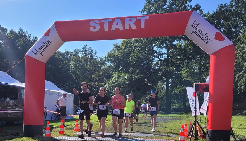 Grupa zawodników przebiega pod dmuchaną brama z napisem "Start". Uczestnicy biegu Ultra Wrocław 3 - Przegoń Raka.
