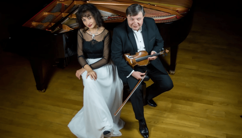 Art Chamber Duo. Kobieta i mężczyzna siedzą obok fortepianu. Mężczyzna trzyma w rękach skrzypce i smyczek.