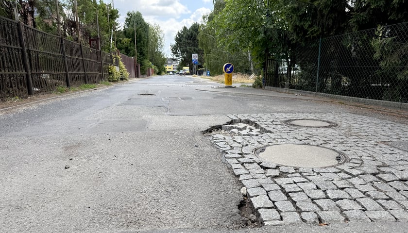 Nawierzchnia ulicy Nefrytowej z wykruszonym asfaltem i poluzowana kostką brukową.