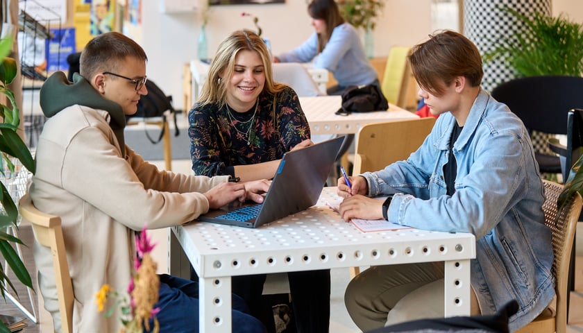 Troje studentów: Jan, Marta i Kacper siedzą przy stoliku kawiarnianym; Jan patrzy w ekran laptopa, Marta rozmawia z Kacprem