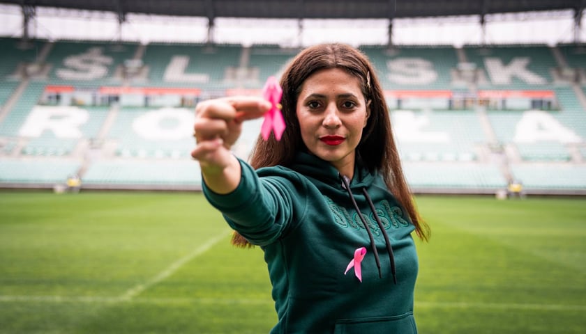 Kobieta z różową wstążeczką na tle stadionu piłkarskiego.