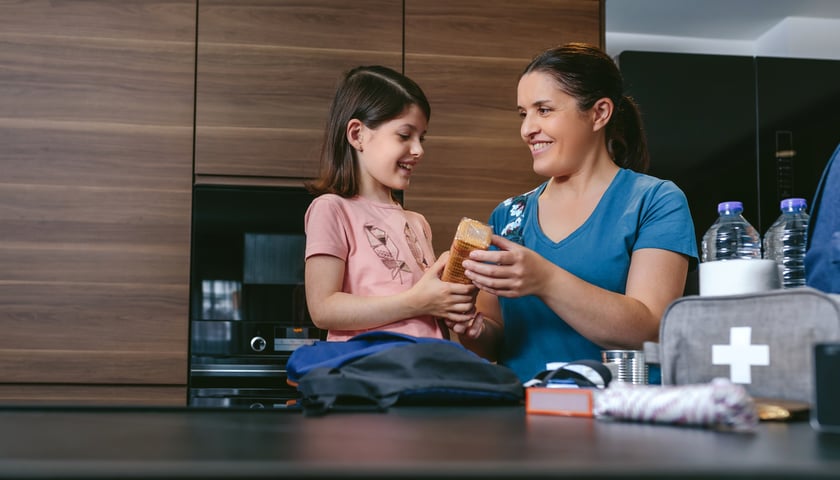 Dziewczynka w różowej koszulce z mamą w niebieskiej bluzce wkładają ubranie do granatowego plecaka
