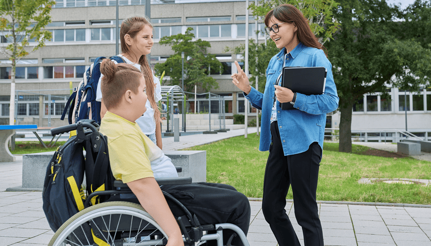Na zdjęciu ilustracyjnym nauczycielka zwracająca się do dwojga dzieci, z których dziewczynka stoi obok chłopca na wózku inwalidzkim