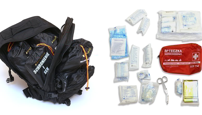 Czarny plecak ewakuacyjny, otwarty, z widocznym czarnym workiem z napisem "Schronienie i sen", a obok czerwone etui apteczki samochodowej, a obok niej rozłożone bandaże i gazy w opakowaniach oraz nożyczki.