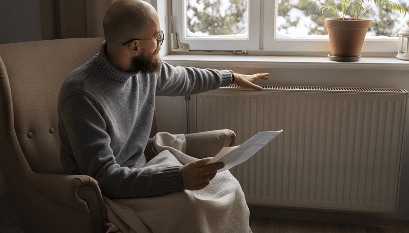 Mężczyzna w okularach, siedzi na fotelu, nogi ma przykryte kocem. W jednej ręce trzyma dokumenty, drugą rękę ma wyciągniętą nad kaloryfer. Zdjęcie ilustracyjne