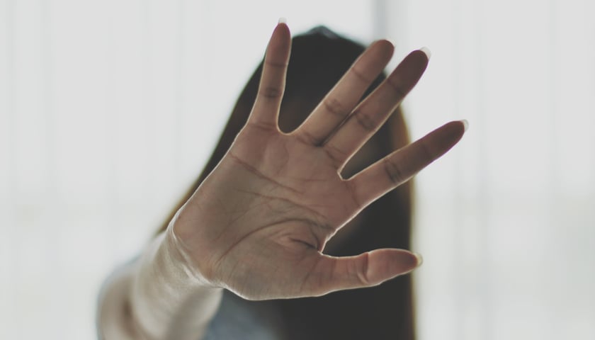 Dłoń z rozłożonymi palcami przed kamerą w geście odmowy lub zatrzymania. Osoba ubrana w jasny materiał, na jasnym tle.