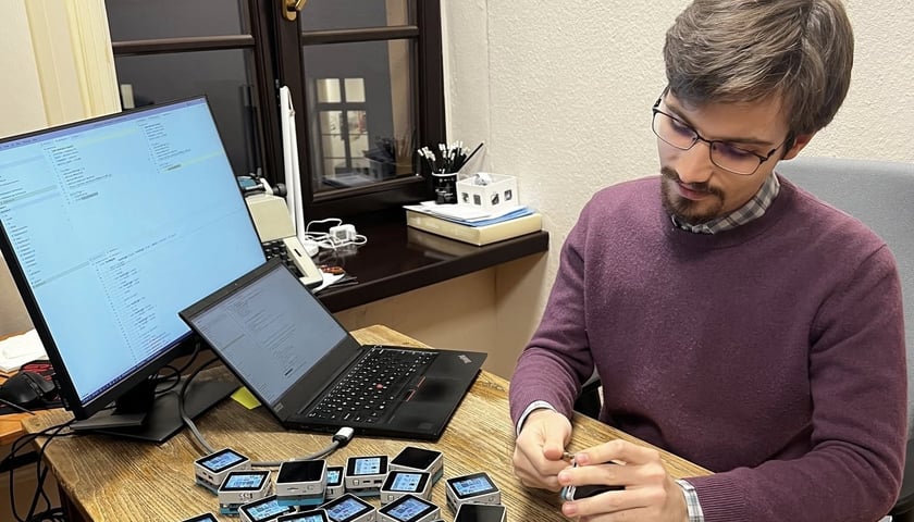 Student siedzi przy biurku przed komputerem i laptopem. Przed nim leżą niewielkie urządzenia do badania.