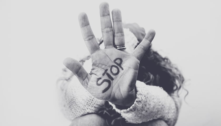 Czarno-białe zdjęcie dłoni osoby z napisem "STOP" na wewnętrznej stronie dłoni. Dłoń jest podniesiona w geście protestu. W tle rozmyty wizerunek osoby. Zdjęcie symbolizuje sprzeciw wobec przemocy.