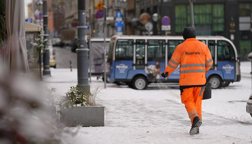 Pierwszy śnieg we Wrocławiu, 24.11.2025. Pracownik z pomarańczowym kombinezonie posypujący solą chodnik.