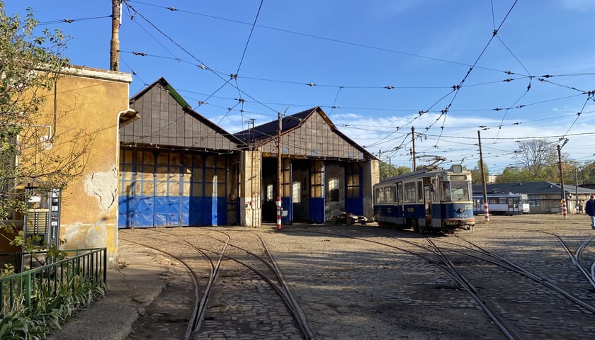 Na zdjęciu znajduje się budynki hal zajezdni z otwartymi bramami. Przed budynkiem na torowisku stojący tramwaj./ The photo shows the depot's hall buildings with their gates open. A tram is parked on the tracks in front of the building.