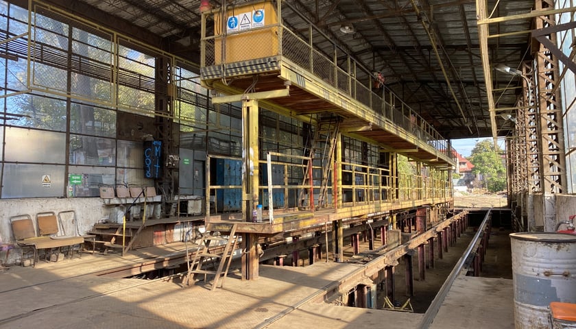 Na zdjęciu znajduje się wnętrze hali zajezdni tramwajowej z dwoma torami oraz pomostem pomiędzy nimi, umożliwiającym prace na dachach tramwajów. Pod torami przestrzeń, dzięki której można zobaczyć podwozia tramwajów./ The photo shows the interior of the tram depot hall, with two tracks and a walkway between them, allowing work to be carried out on the tram roofs. Below the tracks is a space allowing the tram chassis to be seen.