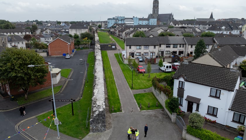 Na zdjęciu widoczne z góry budynki, ulica, chodnik, pojazdy i trawniki. Na środku zdjęcia widoczny mur średniowieczny./ The photo shows buildings, a street, a sidewalk, vehicles, and lawns visible from above. A medieval wall is visible in the center of the photo.