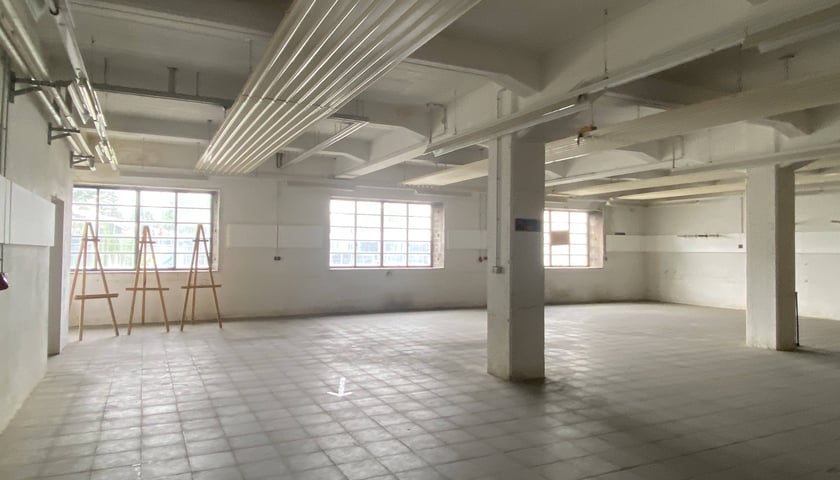 Na zdjęciu widoczne puste wnętrze hali fabryki z dwoma słupami pośrodku. Wnętrze hali oświetlone przez trzy duże okna./ The photo shows the empty interior of the factory hall with two columns in the center. The hall interior is illuminated by three large windows.