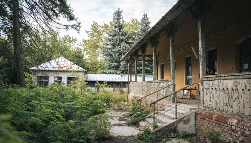 Na zdjęciu widoczne budynki. Bliższy budynek po prawej stronie z podcieniem. Po lewej stronie zdjęcia roślinność, drzewa, niebo z chmurami./ The photo shows buildings. The closer building on the right has an arcade. On the left side of the photo are vegetation, trees, and a sky with clouds.