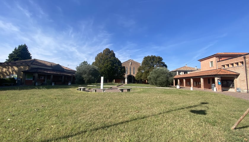 Na zdjęciu widoczny plac pokryty trawą. W jego centralnej części trzy ławki. Plac otoczony trzema niskimi budynkami. W tle drzewa oraz niebo z chmurami./ The photo shows a grass-covered square. Three benches are located in its central area. The square is surrounded by three low-rise buildings. Trees and a cloudy sky are in the background.