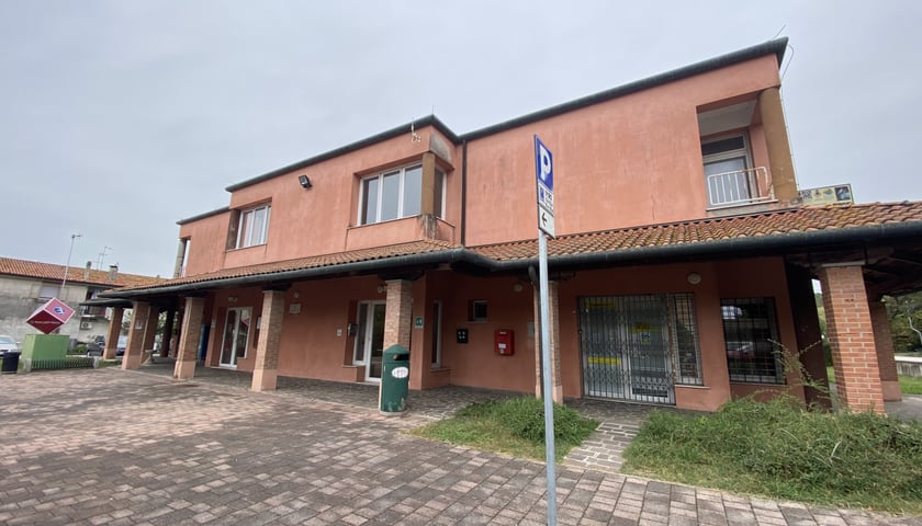 Na zdjęciu widoczny dwukondygnacyjny budynek z podcieniem w części parterowej z kolumnami. Przed budynkiem widoczny chodnik i drogowy znak informacyjny parkowania. W tle pochmurne niebo./ The photo shows a two-story building with a ground-floor arcade and columns. A sidewalk and parking sign are visible in front of the building. A cloudy sky is in the background.