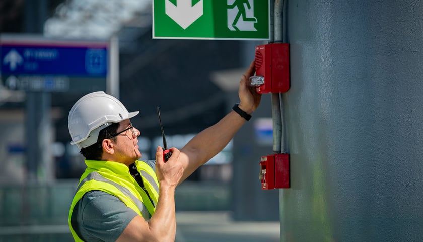 Mężczyzna w kamizelce odblaskowej i kasku budowlanym mówi do krótkofalówki, trzymając rękę na przycisku alarmowym; nad nim biała strzałka na zielonym tle wskazująca drogę ewakuacyjną