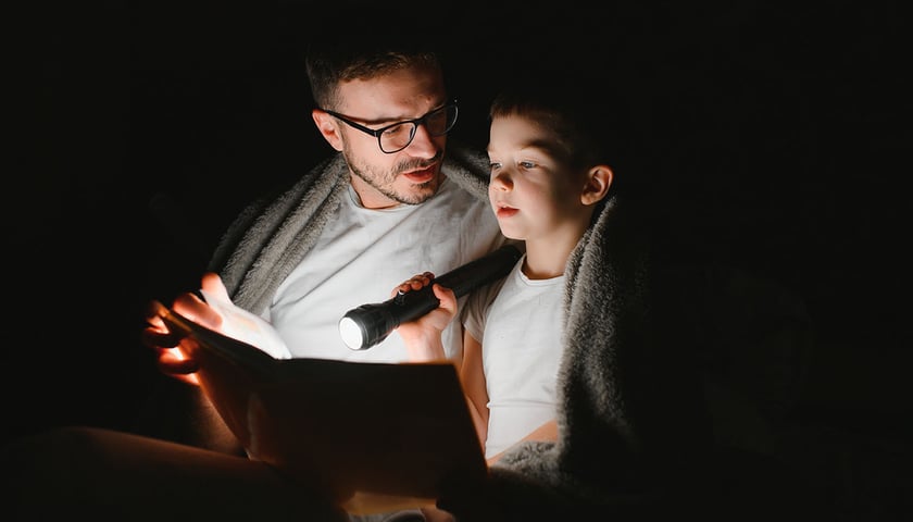 Tata w okularach siedzi z synem nad książką w ciemności - siedzą pod kocem, chłopiec świeci latarką