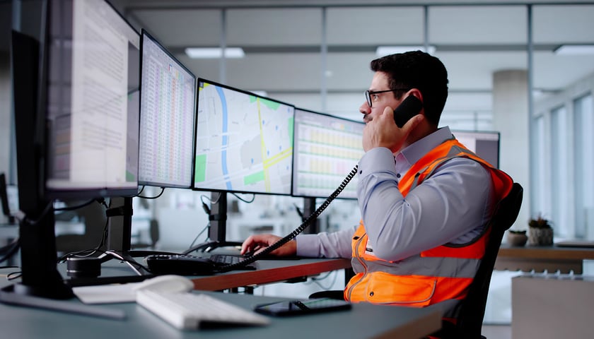 Mężczyzna w okularach, w jasnej koszuli i odblaskowej pomarańczowej kamizelce trzyma słuchawkę telefonu przy uchu i patrzy w ekran komputera