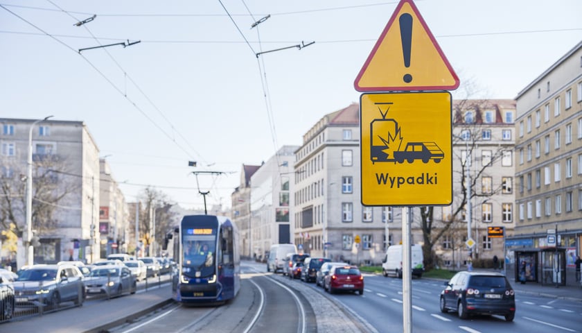 Ulica z torowiskiem i jezdnią oraz widocznym znakiem ostrzegawczym z tablicą zawierającą piktogram kolizji tramwaju z samochodem i napis "Wypadki".  Tablica ostrzegająca o niebezpiecznym miejscu na pl. Legionów.
