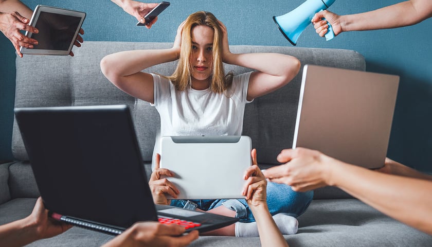 Dziewczyna o blond włosach w białej koszulce i niebieskich dżinsach siedzi po turecku na szarej kanapie, zatykając uszy rękoma. Wokół niej widać ręce, które trzymają: smartfion, tablet, otwarte laptopy i megafon