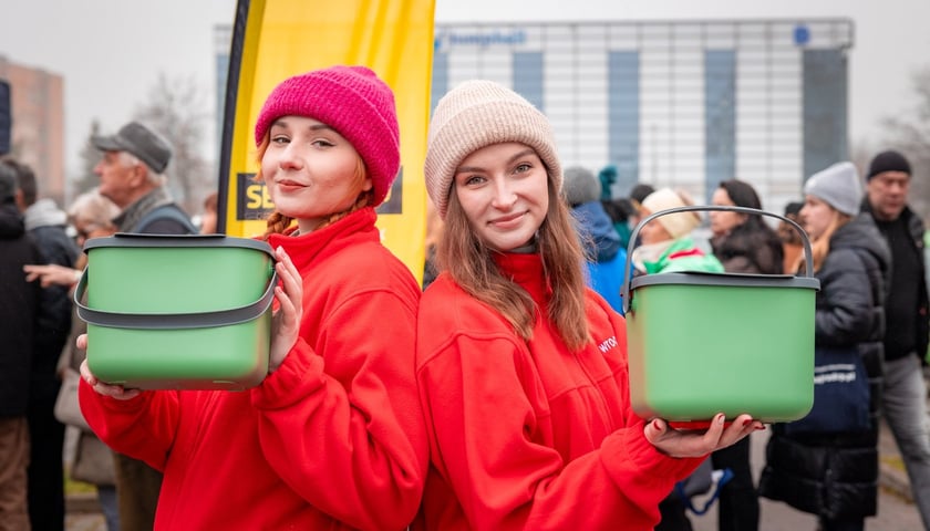 Dwie młode kobiety ubrane w czapki i czerwone bluzy trzymają w rękach pudełka z tworzywa. Akcja dystrybucji pojemników na bioodpady.