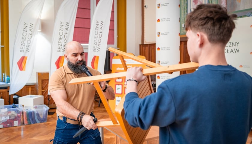 Szkolenie w ramach programu "Bezpieczny Wrocław" w I LO we Wrocławiu. Na zdjęciu mężczyzna z atrapą nożna i chłopak zasłaniający się krzesłem