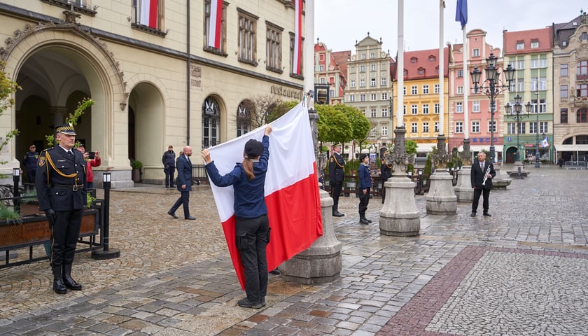 Kobieta w berecie tyłem do obiektywu stoi przed Sukiennicami, trzymając w rękach biało-czerwoną flagę, która zaraz będzie wciągana na maszt