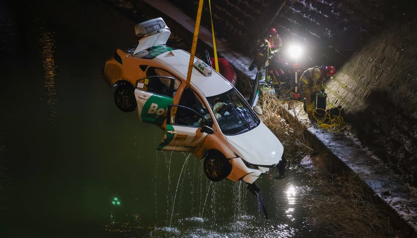 Samochód zawieszony na taśmach i wyciągany z Odry przez dźwig. W tle na nabrzeżu strażacy. Wieczorna akcja służb ratowniczych po wpadnięciu auta osobowego do Odry przy Kępie Mieszczańskiej 4 stycznia 2026 r.