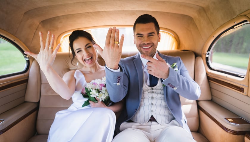 Na zdjęciu młoda para we wnętrzu luksusowego samochodu pokazuje obrączki na dłoniach. Zdjęcie ilustracyjne