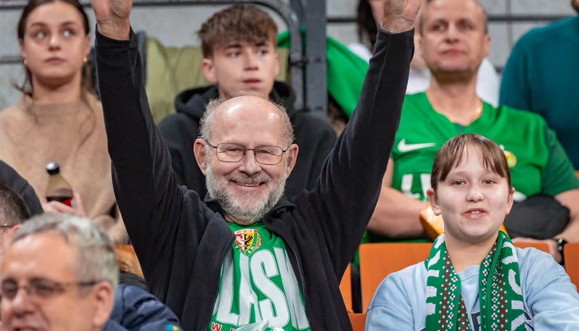 Uśmiechnięty mężczyzna w zielonej koszulce Śląska i bluzie, obok niego kobieta i inni kibice na trybunie Hali Stulecia na meczu z Arisem Saloniki, 20 stycznia 2026. 