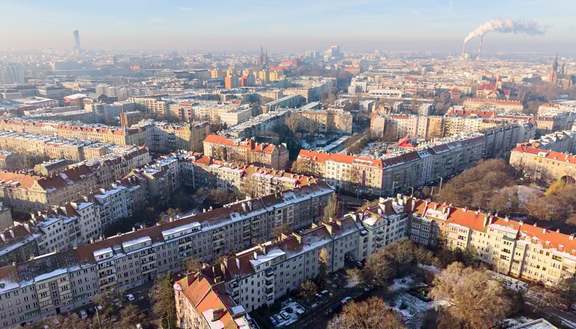 Zdjęcie z lotu ptaka przedstawiające w zimowy słoneczny dzień ulice zabudowane kamienicami z widokiem na podwórka i panoramę  miasta.