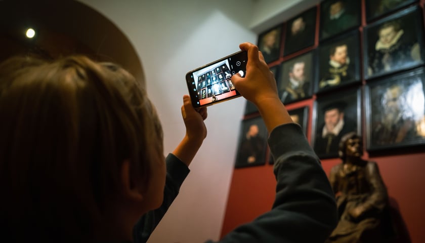 Osoba zwiedzająca muzeum robi telefonem zdjęcie obrazów na ścianie.