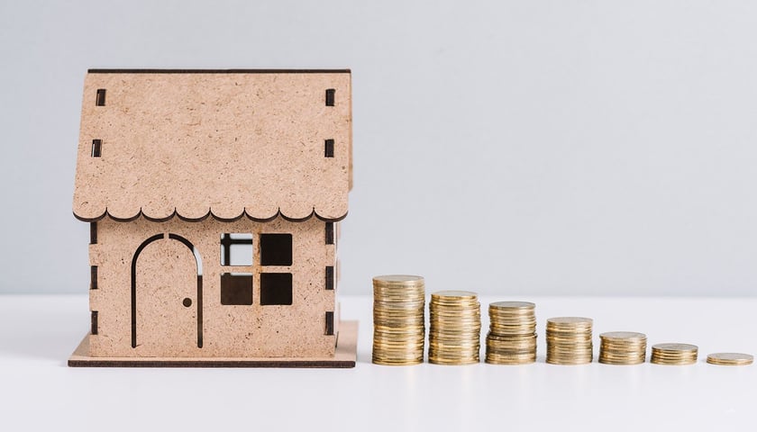 Zdjęcie ilustracyjne przedstawia makietę domu i stosy monet
