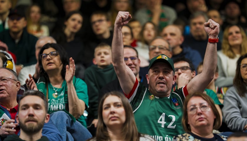 Kibic w zielonej koszulce Śląska i w czapce z daszkiem, unoszący ręce na trybunie Hali Orbita podczas spotkania z Gliwicami. Na zdjęciu są także inni kibice.