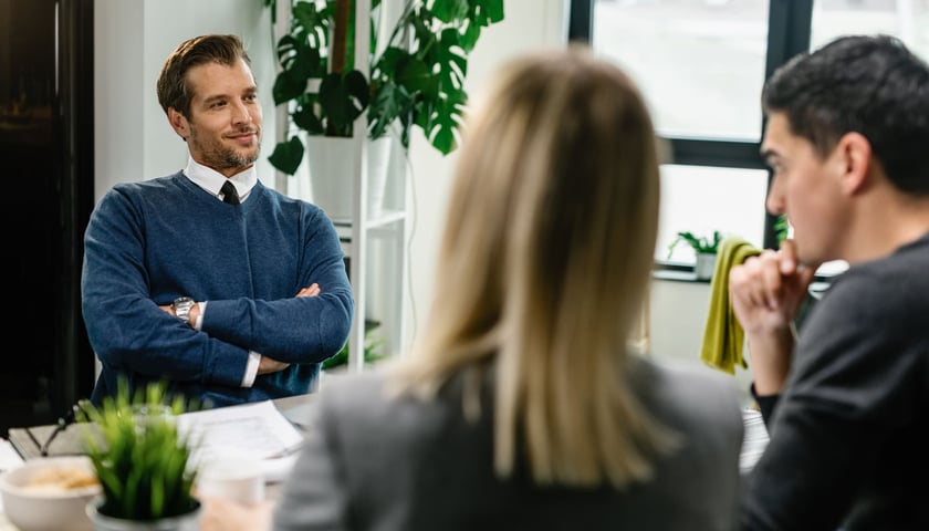 Negocjacje przy stole. Mężczyzna z założonymi rękami siedzi naprzeciwko mężczyzny i kobiety, których widzimy od tyłu. Zdjęcie ilustracyjne.